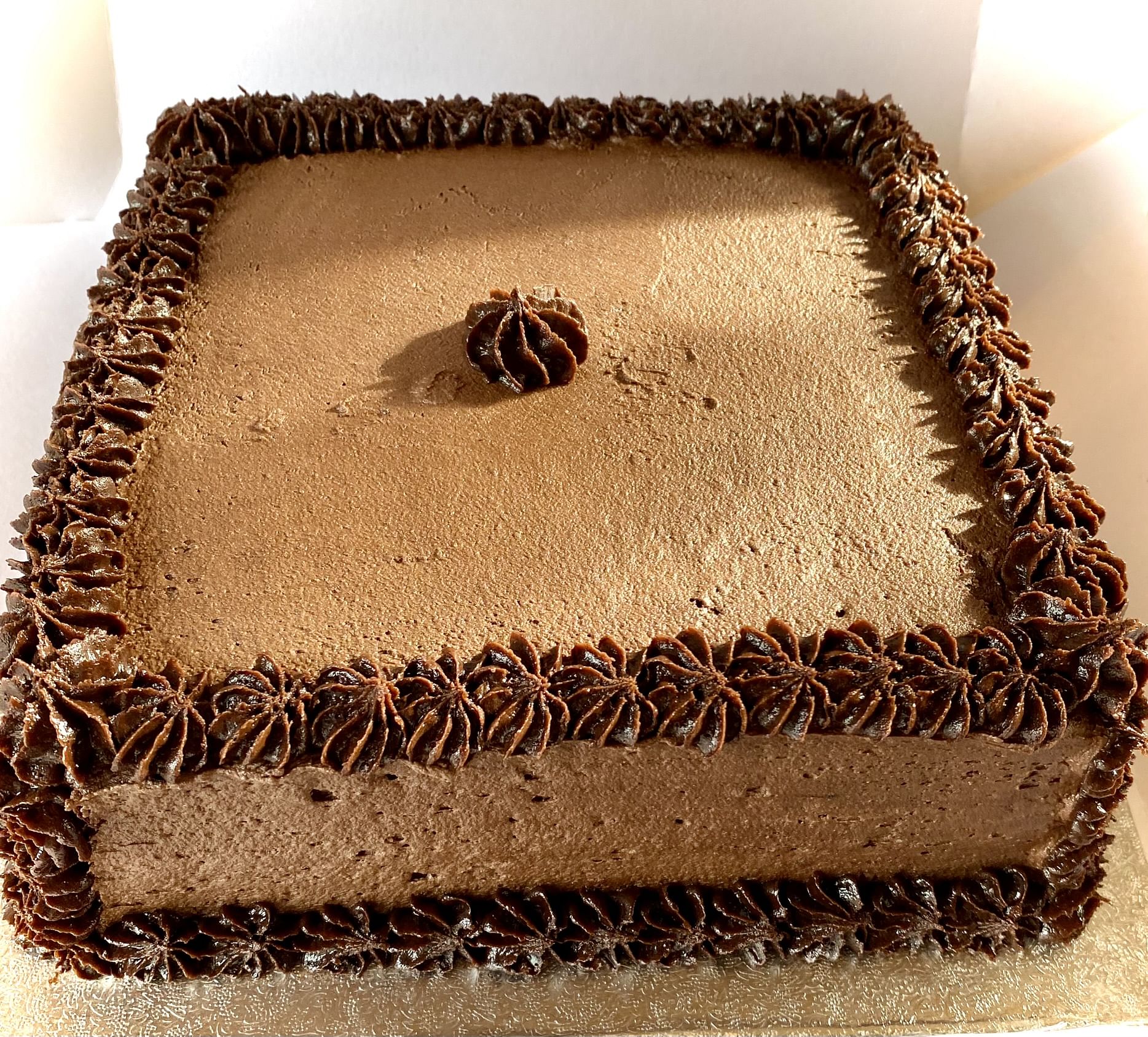 A square chocolate cake adorned with a border of chocolate frosting rosettes along the top edge and a single rosette in the center. This decadent dessert is elegantly placed on a golden base against a white background.